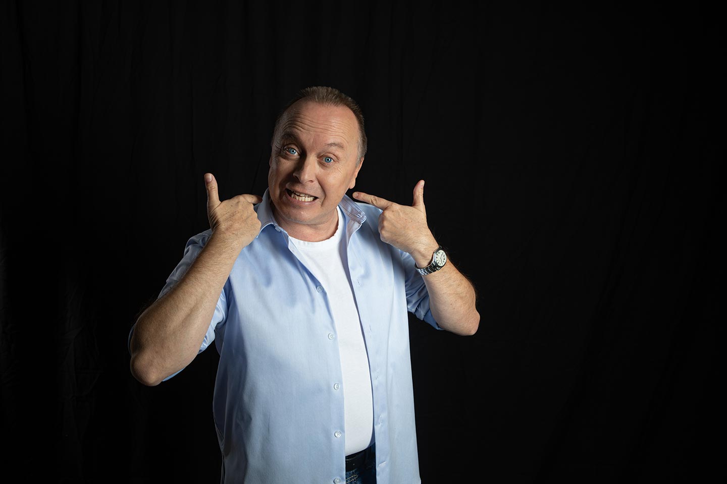 Christoph Rosenberger zeigt mit beiden Daumen auf sich selbst und blickt lebhaft in die Kamera vor dunklem Hintergrund.