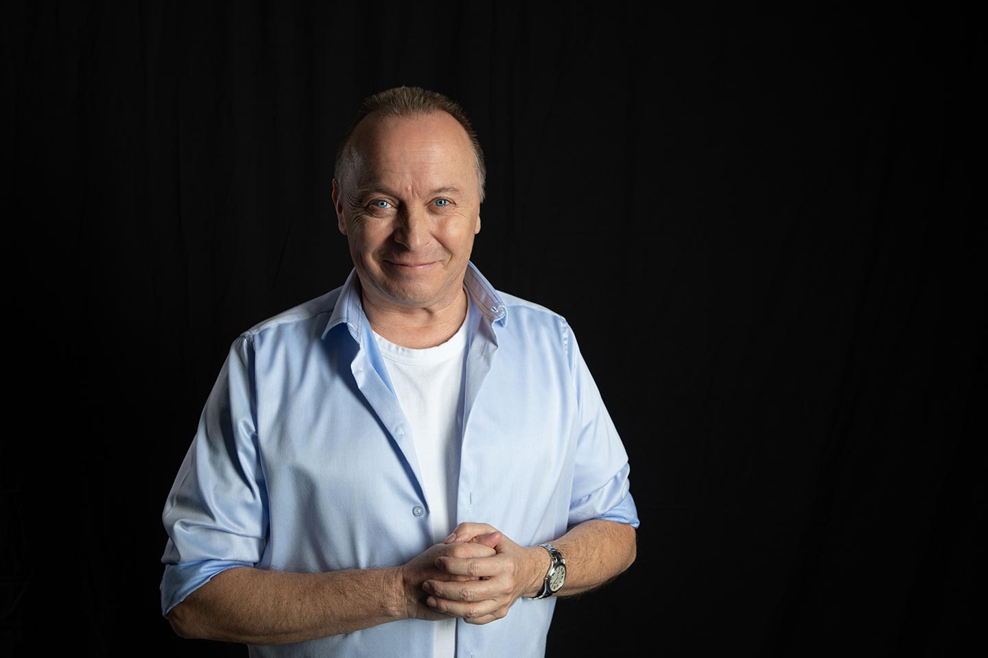 Christoph Rosenberger steht mit gefalteten Händen und leichtem Lächeln vor dunklem Hintergrund.