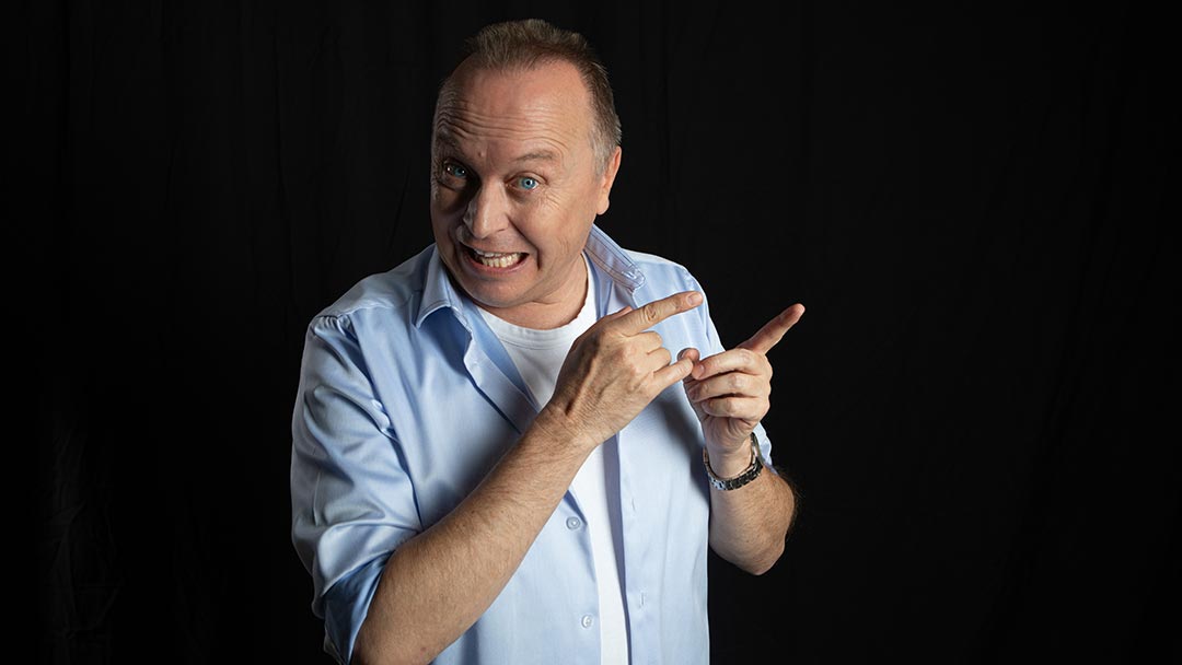 Christoph Rosenberger mit aufgewecktem Gesichtsausdruck zeigt mit beiden Händen nach rechts vor dunklem Hintergrund.