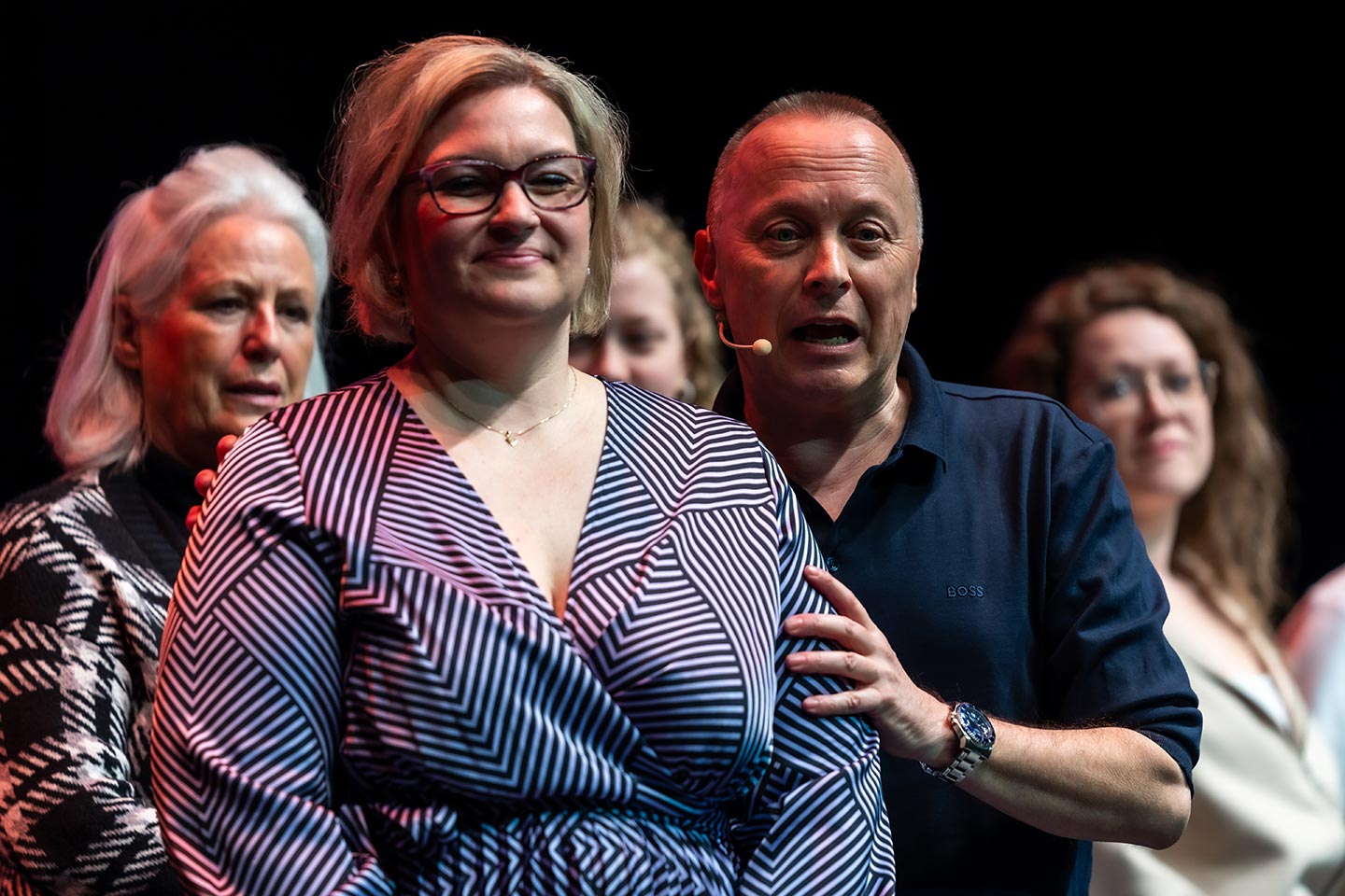 Mehrere Personen stehen auf einer Bühne. Im Vordergrund steht eine Person, dahinter spricht Christoph Rosenberger mit Headset und legt ihr die Hand auf die Schulter; weitere Personen stehen im Hintergrund.