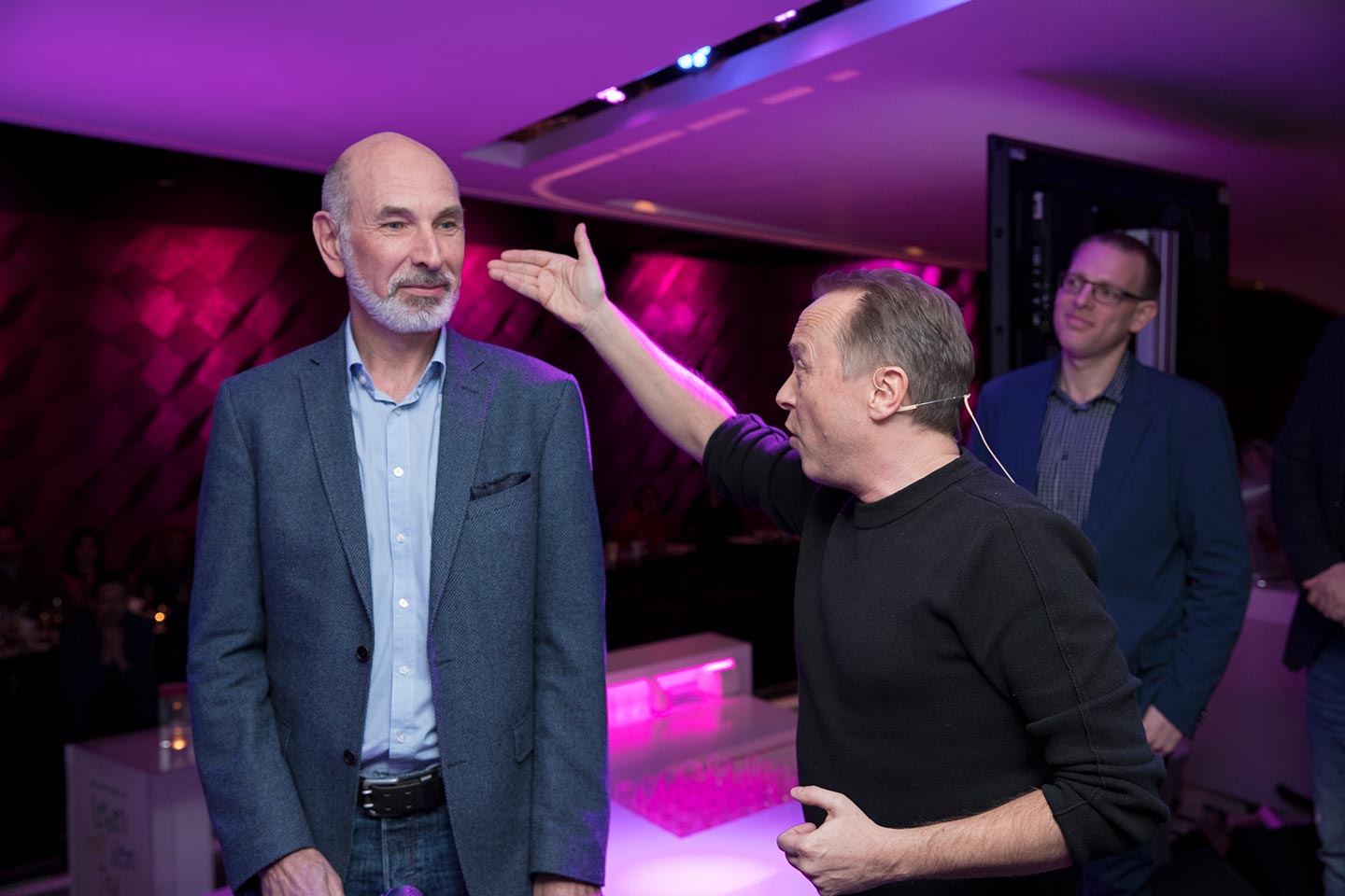 Mehrere Personen stehen in einem Veranstaltungsraum mit farbigem Licht. Christoph Rosenberger mit Headset erklärt etwas und hebt die Hand neben dem Kopf einer anderen Person; weitere Personen stehen beobachtend im Hintergrund.